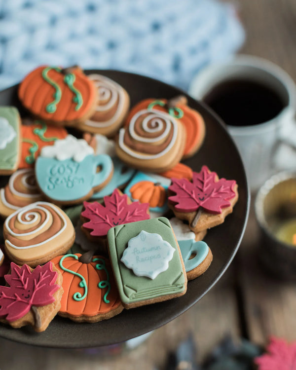 Honeywell Bakes The Cosy Biscuit Jar - A plate of decorated autumn-themed sugar cookies, including pumpkins, fall leaves, a mug cookie that says 'Cute Snaps', and swirls, styled on a dark plate with a cup of coffee in the background.