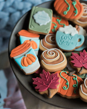 Honeywell Bakes The Cosy Biscuit Jar - A close-up of assorted decorated cookies on a dark plate, including pumpkin-shaped cookies, fall leaves, a coffee cup cookie with ‘Cosy Season' and whipped cream, cinnamon swirls, and other autumn-themed designs.