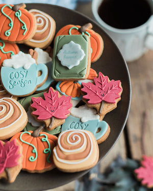 Honeywell Bakes The Cosy Biscuit Jar - A close-up of an assortment of decorated autumn-themed sugar cookies arranged on a dark plate, including pumpkin, maple leaf, mug, and book-shaped cookies, with a coffee cup blurred in the background.