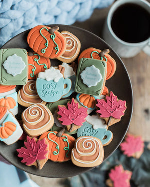 Honeywell Bakes The Cosy Biscuit Jar - A plate of decorated autumn-themed sugar cookies, including pumpkin shapes, fall leaves, cinnamon swirls, and mug designs with the words 'Cosy Season', shown on a dark plate with a coffee mug in the background.