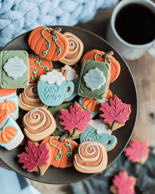 Honeywell Bakes The Cosy Biscuit Jar - A plate of decorated autumn-themed sugar cookies, including pumpkin shapes, fall leaves, cinnamon swirls, and mug designs with the words 'Cosy Season', shown on a dark plate with a coffee mug in the background.