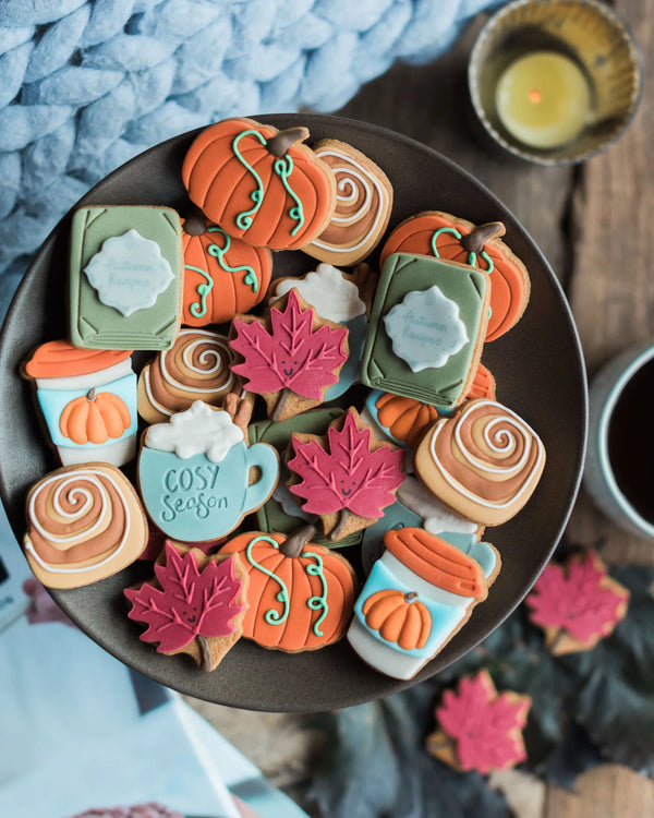 Honeywell Bakes The Cosy Biscuit Jar - An overhead styled shot of a dark plate filled with decorated autumn-themed cookies, including pumpkin shapes, fall leaves, cinnamon swirls, and a mug cookie that says 'Cosy Season,' set on a rustic wooden table with a blue knit blanket and candle in the background.