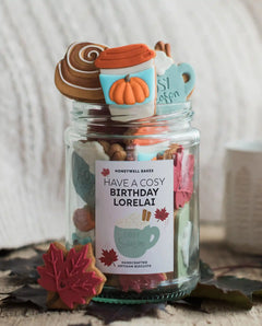 Honeywell Bakes The Cosy Biscuit Jar - A clear glass jar filled with assorted baked goods and topped with fall-themed treats, including a pumpkin cup decoration, a cinnamon swirl, and a mug topper. The jar has a label that reads 'HAVE A COSY BIRTHDAY LORELAI' and 'HONEYWELL BAKES' at the top.