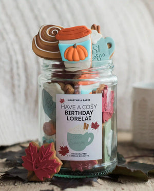 Honeywell Bakes The Cosy Biscuit Jar - A clear glass jar filled with assorted baked goods and topped with fall-themed treats, including a pumpkin cup decoration, a cinnamon swirl, and a mug topper. The jar has a label that reads 'HAVE A COSY BIRTHDAY LORELAI' and 'HONEYWELL BAKES' at the top.