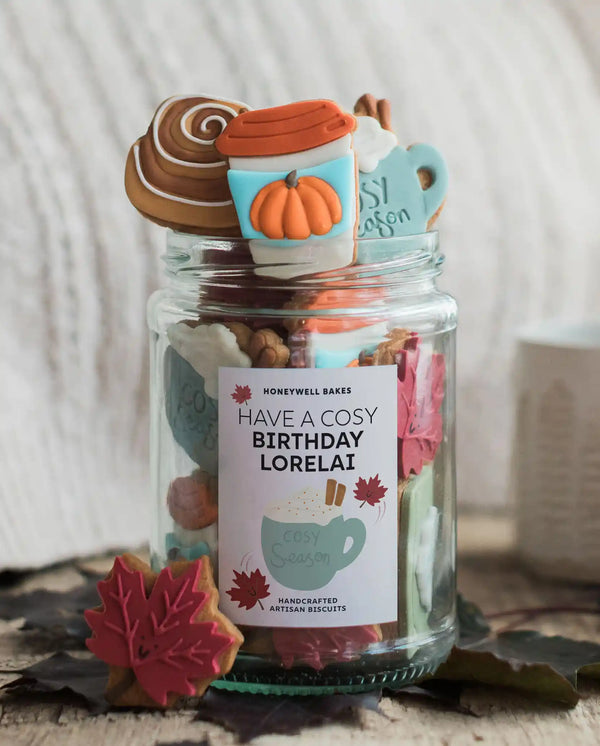 Honeywell Bakes The Cosy Biscuit Jar - A clear glass jar filled with assorted baked goods and topped with fall-themed treats, including a pumpkin cup decoration, a cinnamon swirl, and a mug topper. The jar has a label that reads 'HAVE A COSY BIRTHDAY LORELAI' and 'HONEYWELL BAKES' at the top.