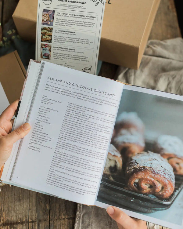 Master Bakers Bundle - An open cookbook showing a recipe page for almond and chocolate croissants, held in hands over a rustic wooden table. A brown cardboard box and another book are visible in the background.