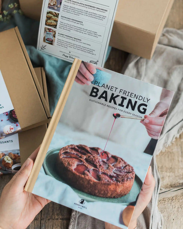 Master Bakers Bundle A person holds a hardcover book titled 'Planet Friendly Baking' with a wooden spine, showing a baked cake on the cover. The scene is styled with other baking books and cardboard boxes in the background.