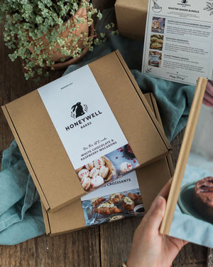 Master Bakers Bundle - A Honeywell Bakes gift box set on a rustic wooden table, styled with a potted trailing plant, teal fabric, and a hand holding a recipe booklet.