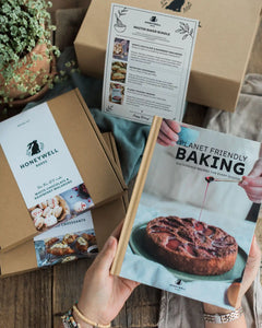 Master Bakers Bundle - A person holds a copy of the Honeywell Baking 'Planet Friendly Baking' cookbook in one hand while another hand points to the cover. Several brown cardboard Honeywell Baking boxes are stacked in the background on a rustic wooden table next to a potted plant.