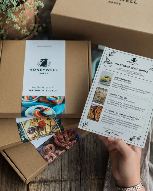 Plant Based Bread Bundle - A hand holds a printed recipe card for Honeywell Bakes Rainbow Bagels over an open kraft paper box, with another branded box and recipe cards visible on a wooden tabletop.