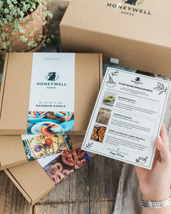 Plant Based Bread Bundle - A Honeywell Bakes gift box set displayed on a rustic wooden table, featuring a large brown kraft box, a smaller box with a white label reading Rainbow Bagels, and a hand holding a printed recipe card for Plant Based Bread Bundle.