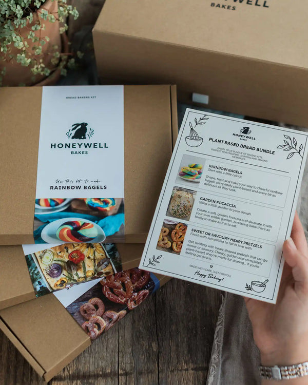 Plant Based Bread Bundle - A Honeywell Bakes gift box set on a rustic wooden table, with a hand holding a printed recipe card for Rainbow Bagels. The set includes a kraft paper box with a white Honeywell label, and recipe cards.