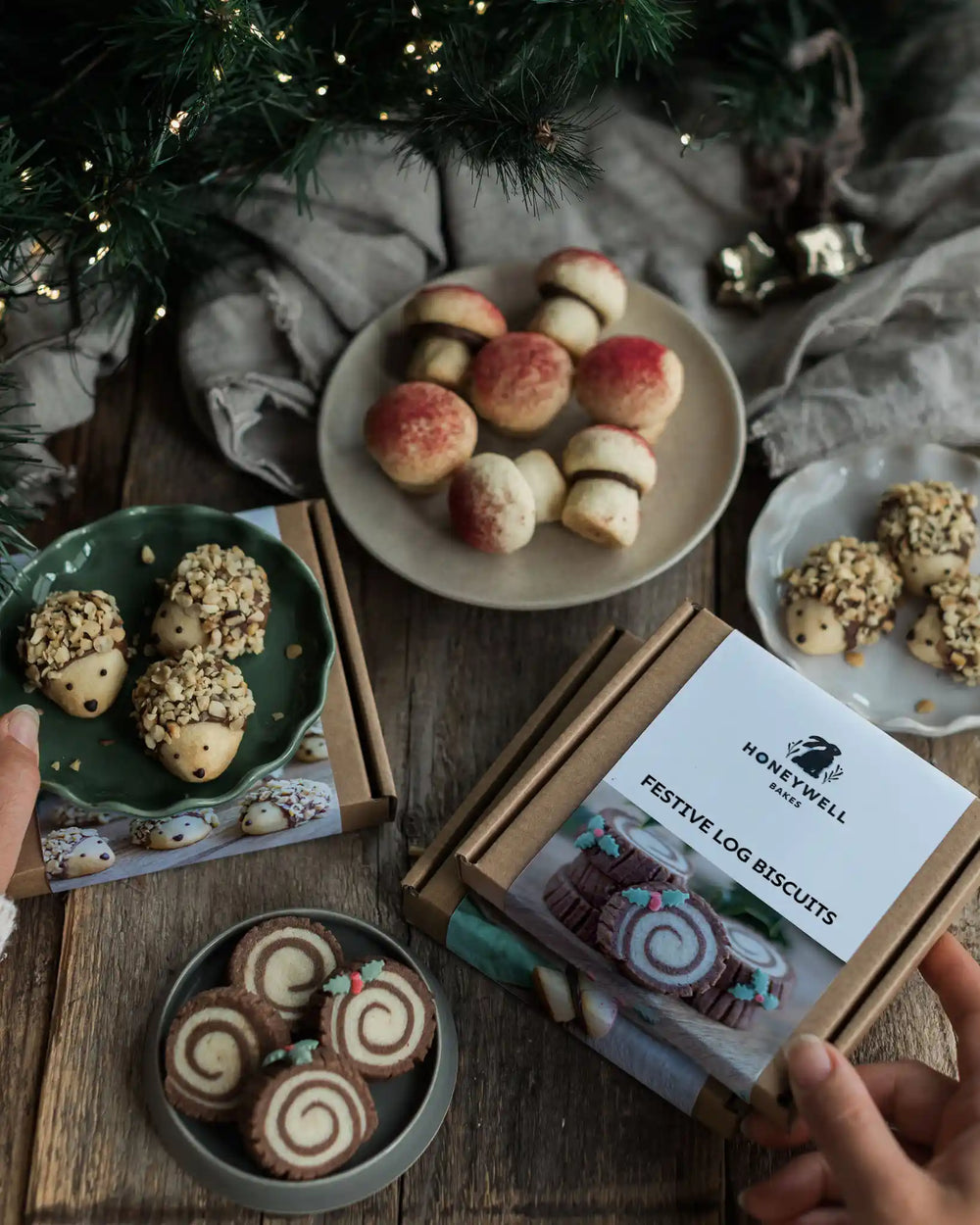 The Woodland Baking Kit Collection - A styled flat-lay of Honeywell Bakes 'Festive Log Biscuits' in a kraft paper box, surrounded by plates of decorated cookies and festive holiday decorations on a rustic wooden table.