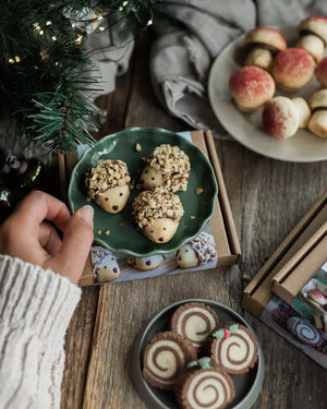 The Woodland Baking Kit Collection - A styled holiday tabletop scene showing decorated acorn-shaped cookies on a green plate, with a hand reaching in from the left. Other plates of cookies, gift boxes, pine branches, and a gray cloth are arranged on a wooden table.