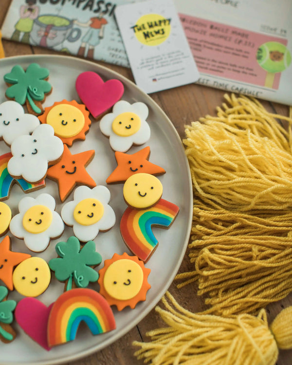 Honeywell Bakes The Happy News Biscuit Jar - Colourful cookies shaped like smiley faces, rainbows, and hearts on a plate with yellow yarn.