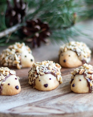 The Woodland Baking Kit Collection - Five small baked cookies shaped like hedgehogs, topped with chopped nuts and dark chocolate, arranged on a wooden surface with pine branches and a pinecone in the blurred background.