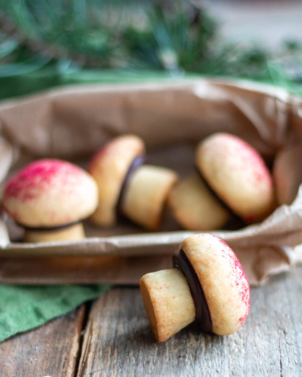 The Woodland Baking Kit Collection - Small cookies shaped like mushrooms, with tan cookie bodies, dark chocolate stems, and red sprinkles on the caps, displayed on a rustic wooden table with a brown paper bag in the background.