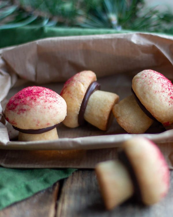 The Woodland Baking Kit Collection - Mushroom-shaped cookies with red sprinkles, arranged in a brown paper-lined dish on a rustic wooden surface with green pine branches in the background.