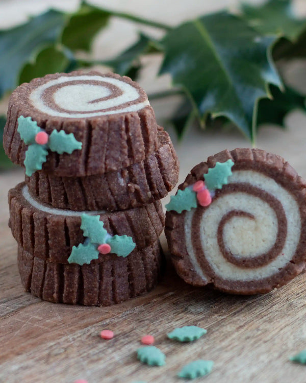 The Woodland Baking Kit Collection - Decorated Christmas log cookies stacked on a wooden surface with holly leaves in the background. The cookies have a dark brown and light cream swirl pattern and are adorned with teal holly leaves and small pink berries.