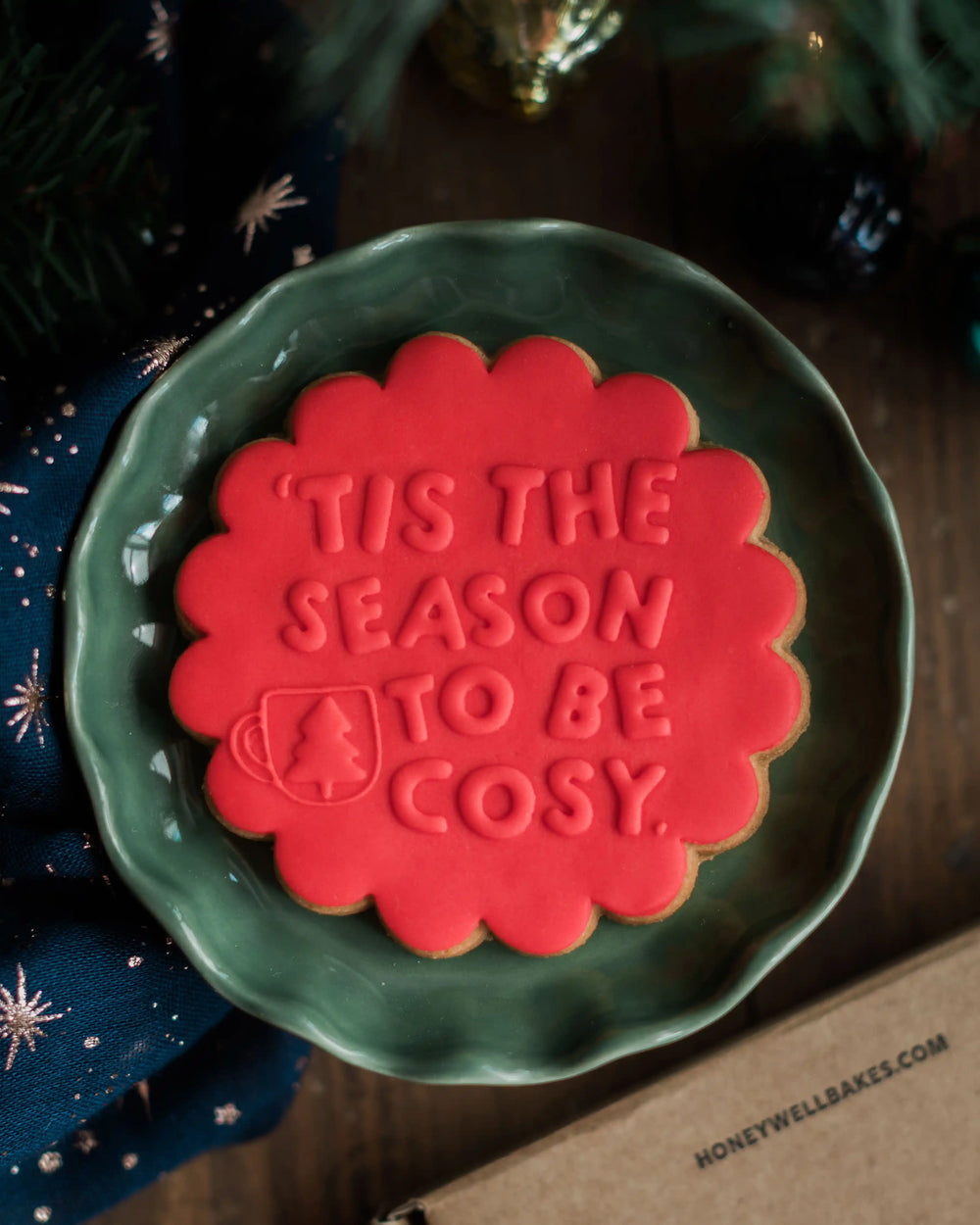 Honeywell Bakes Tis The Season Biscuit - Red cookie with 'Tis the Season to be Cosy' text on a green plate, Christmas tree in background