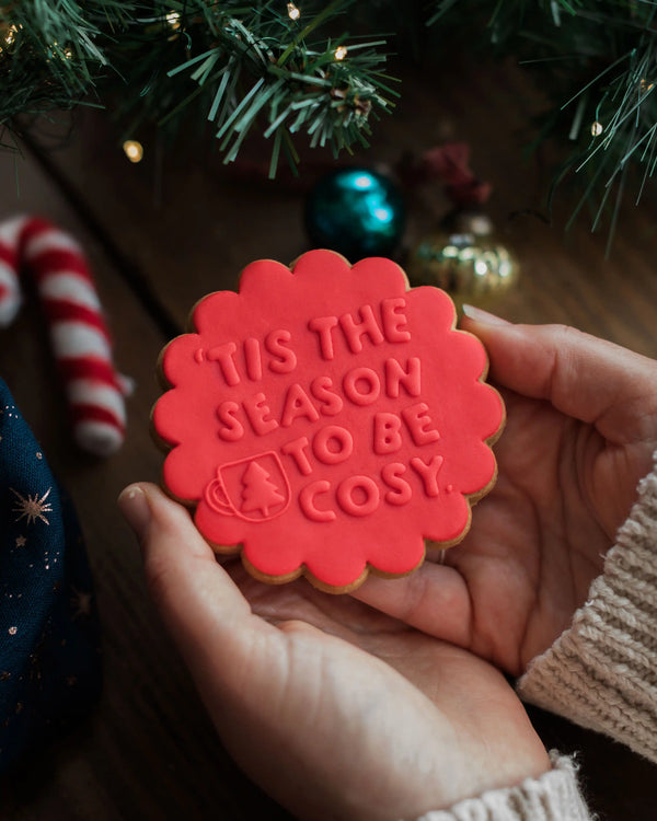 Honeywell Bakes Tis The Season Biscuit - Red cookie with 'Tis the season to be cosy' text held by a person in front of a Christmas tree.