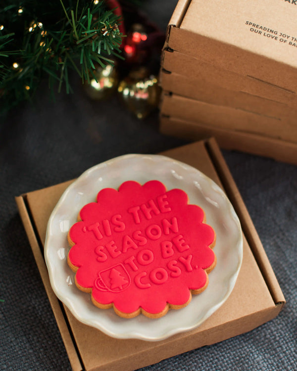 Honeywell Bakes Tis The Season Biscuit x 5 - Red cookie with 'Tis the season to be cozy' text on a white plate, placed in a brown box with Christmas decorations in the background.