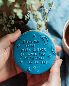 Honeywell Bakes To The Moon & Back - Blue cookie with 'I love you to the moon and back' text held by a hand, with a plant and cup in the background.