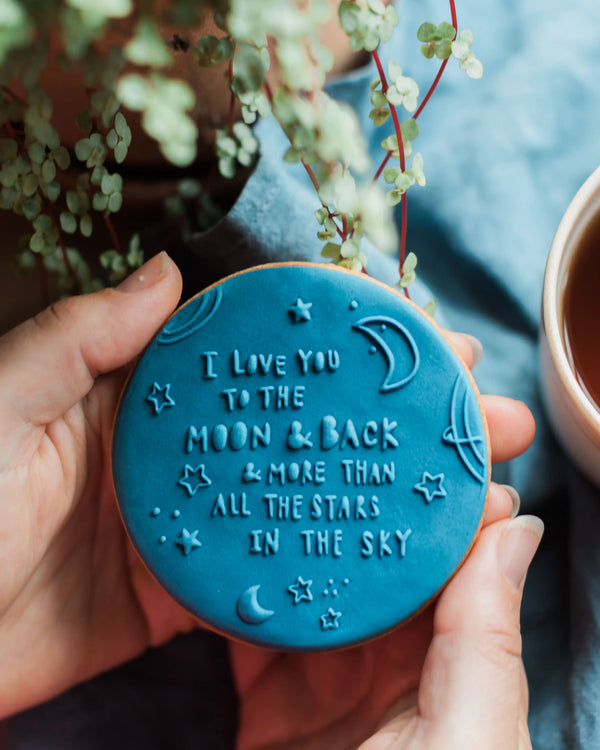 Honeywell Bakes To The Moon & Back - Blue cookie with 'I love you to the moon and back' text held by a hand, with a plant and cup in the background.
