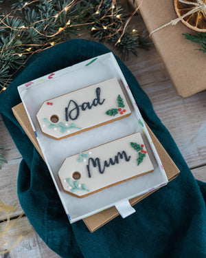 Honeywell Bakes Two Personalised Christmas Tag Biscuits - Decorative cookies shaped like gift tags with 'Dad' and 'Mum' on a wooden surface with greenery and a gift box.