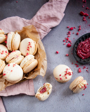 Honeywell Bakes White Chocolate & Raspberry Macaron Kit - White macarons with raspberry pieces and white chocolate drizzle on a grey surface with a pink cloth and raspberry puree.