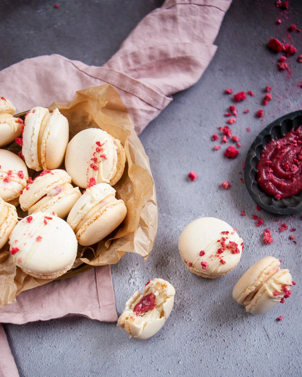Honeywell Bakes White Chocolate & Raspberry Macaron Kit - White macarons with raspberry pieces and white chocolate drizzle on a grey surface with a pink cloth and raspberry puree.