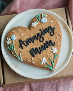 Honeywell Bakes Wildflower Heart – Piped Personalised Biscuit - Heart-shaped cookie with 'Amazing Mum' text on a white plate