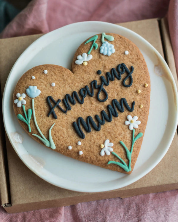 Honeywell Bakes Wildflower Heart – Piped Personalised Biscuit - Heart-shaped cookie with 'Amazing Mum' text on a white plate