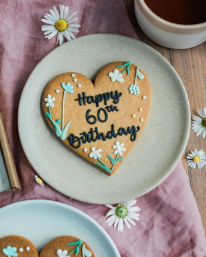 Honeywell Bakes Wildflower Heart – Piped Personalised Biscuit - Heart-shaped cookie with 'Happy 60th Birthday' on a plate with flowers and tea.