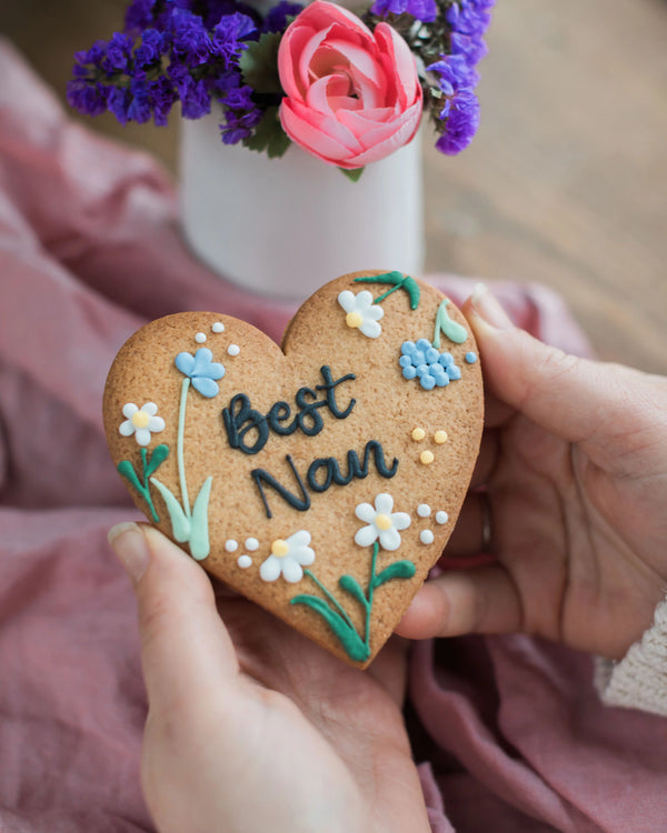 Honeywell Bakes Wildflower Heart – Piped Personalised Biscuit - Heart-shaped cookie with 'Best Nan' text and floral decorations held by a person, with flowers in the background.