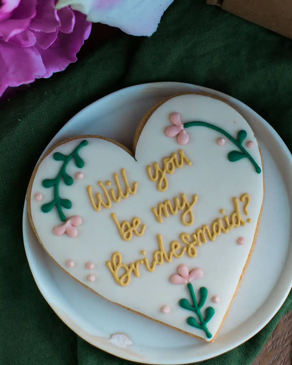 Honeywell Bakes Will You Be My Bridesmaid Biscuit - Heart-shaped cookie with 'Will you be my Bridesmaid?' text on a white plate with flowers in the background.