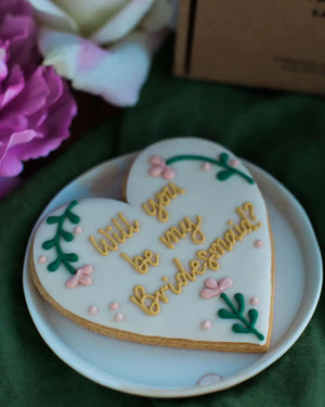 Honeywell Bakes Will You Be My Bridesmaid Biscuit - Heart-shaped cookie with 'Will you be my bridesmaid?' text on a white plate with flowers in the background