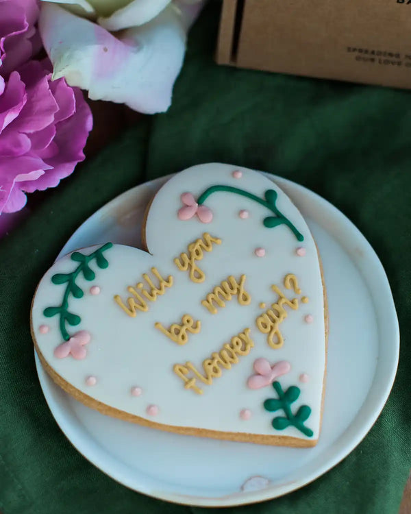 Honeywell Bakes Will You Be My Flower Girl Biscuit - Heart-shaped cookie with 'Will you be my flower girl?' text on a white plate with flowers in the background.