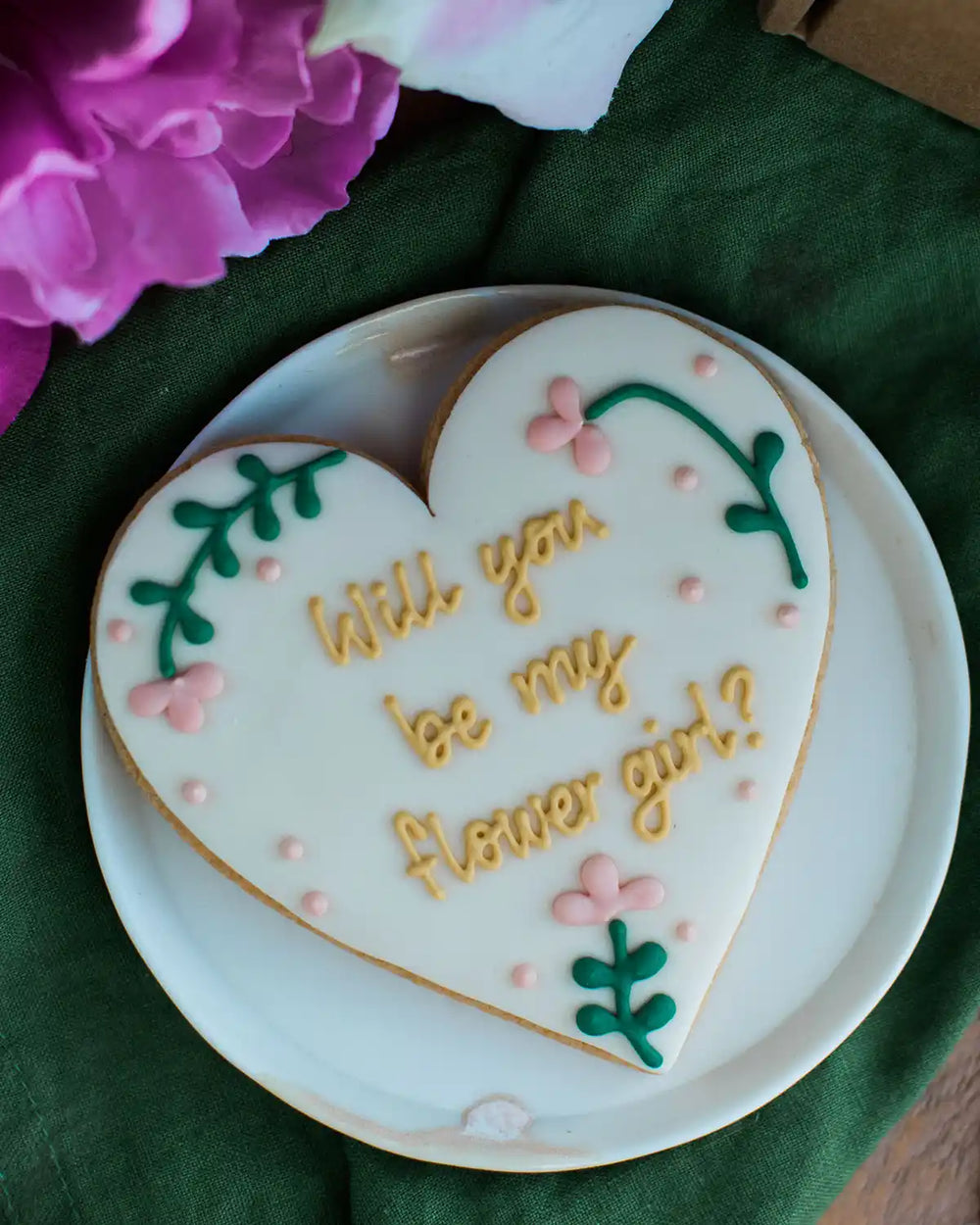 Honeywell Bakes Will You Be My Flower Girl Biscuit - Heart-shaped cookie with 'Will you be my flower girl?' text on a white plate with flowers in the background.