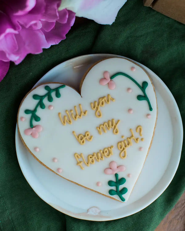 Honeywell Bakes Will You Be My Flower Girl Biscuit - Heart-shaped cookie with 'Will you be my flower girl?' text on a white plate with flowers in the background.