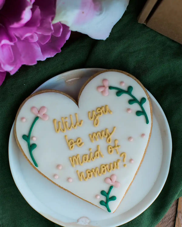 Honeywell Bakes Will You Be My Maid Of Honour Biscuit - Heart-shaped cookie with 'Will you be my Maid of Honour?' text on a white plate with flowers in the background.