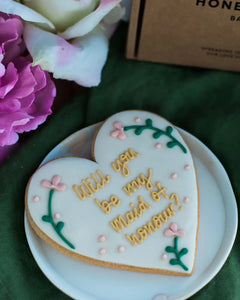 Honeywell Bakes Will You Be My Maid Of Honour Biscuit - Heart-shaped cookie with 'Will you be my Maid of Honour?' text on a white plate with flowers in the background.