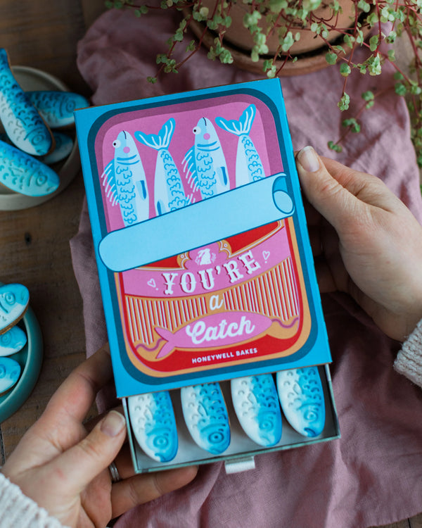 Honeywell Bakes You're a Catch Biscuit Gift Set - packaging with blue sardine fish-shaped hand iced vanilla cookies on a wooden table.